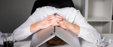 A man with his face pressing against the table looking stressed, with his laptop folded over his head