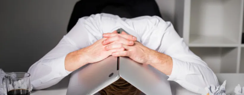 A man with his head buried into his desk looking defeated with his laptop over his head