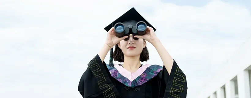 A recent university graduate in her robe looking through binoculars