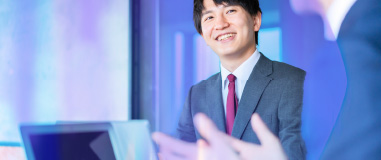 スーツを着た男性が会議中に笑顔を見せているビジネスシーン。手前には同僚のジェスチャーが見え、明るく前向きな職場の雰囲気が伝わる。